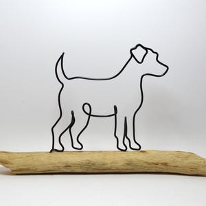 May include: Wire sculpture of a dog standing on a piece of driftwood. The dog is made of black wire and is in a profile pose. The driftwood is light brown and has a natural, rough texture.