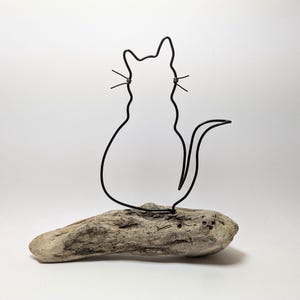 May include: A black wire cat sculpture sitting on a piece of driftwood. The cat is depicted in a simple outline, with whiskers. The driftwood is a light brown color and has a natural, weathered appearance. The sculpture is set against a white background.