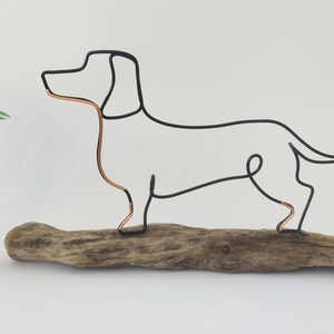 May include: Wire sculpture of a dachshund dog standing on a piece of driftwood. The dog is made of black wire with copper accents.