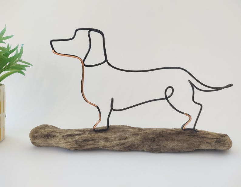 May include: Wire sculpture of a dachshund dog standing on a piece of driftwood. The dog is made of black wire with copper accents.