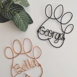 May include: Two wire paw print ornaments shaped from metal wire. One ornament is black wire with the name George formed in cursive script, and the other is copper-coloured wire with the name Bella formed in cursive script within the paw shape.