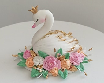 May include: A white fondant swan cake topper with a gold crown, surrounded by pink, peach, and white flowers and green leaves.