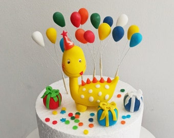 May include: A white cake decorated with a yellow dinosaur wearing a party hat, surrounded by colorful balloons and presents. The cake is also decorated with sprinkles.