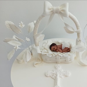 Könnte beinhalten: Weißer Fondantkuchen, verziert mit einer weißen Babyfigur in einem Korb, einem weißen Kreuz und weißen Blumen.