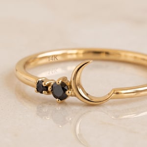 May include: A gold ring featuring a crescent moon shape and two small round black stones. The interior of the band is clearly engraved with the text 14K, indicating the metal purity of this piece of jewellery.