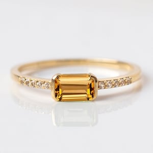 Puede incluir: Un anillo de oro con una piedra preciosa rectangular de color amarillo dorado. La fina banda de oro tiene pequeñas piedras transparentes a los lados de la piedra principal. El anillo se muestra sobre una superficie blanca, reflejando la luz.