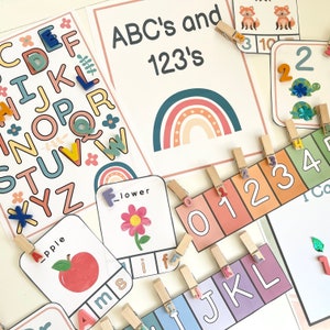 May include: A set of colorful alphabet and number flashcards with rainbow and flower illustrations. The cards are designed for children to learn the alphabet and numbers. The cards are arranged on a white surface with wooden clothespins.