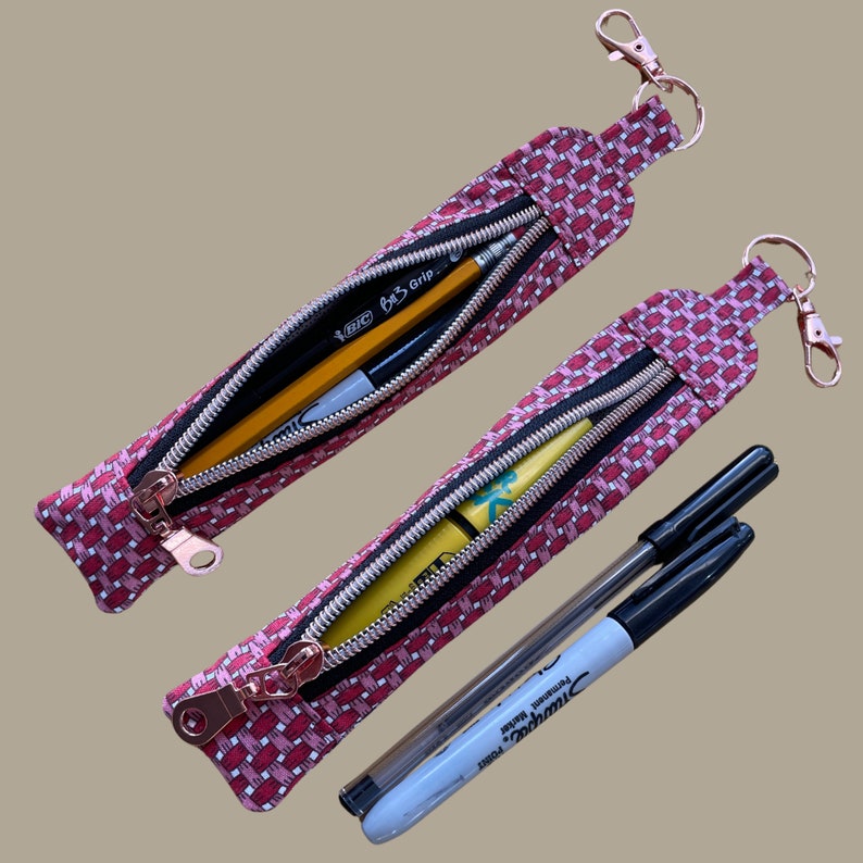 May include: Two pink zippered pouches with keychains. The pouches are filled with pens, pencils, and highlighters. The pouches are made of a patterned fabric with a pink and white geometric design.