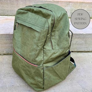 May include: A green backpack with a front pocket and a side pocket. The backpack has a zip closure and a pink zip pull. The text "PDF SEWING PATTERN" is in a circle on the right side of the backpack.