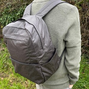 May include: A dark grey backpack with a rectangular design and several zipped compartments. The backpack features adjustable shoulder straps and a top handle. The material looks water-resistant. The person in the image is wearing a green jumper.