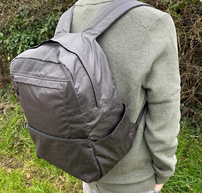 May include: A dark grey backpack with a rectangular design and several zipped compartments. The backpack features adjustable shoulder straps and a top handle. The material looks water-resistant. The person in the image is wearing a green jumper.