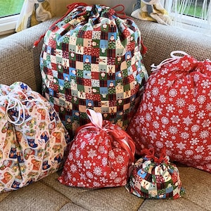 May include: Five drawstring fabric gift bags in various sizes and patterns. The bags feature festive designs, including a patchwork pattern with holiday motifs, a red snowflake print, and a print with Christmas stockings and other holiday elements. The bags are tied with drawstrings.