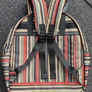 May include: Backpack featuring vertical stripes in red, green, tan, black, and white. It has black adjustable straps and a black handle. The straps are connected with a black buckle. The backpack is on a grey surface.