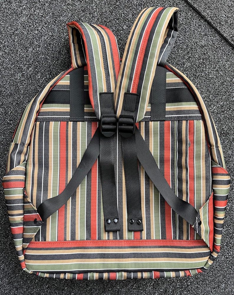 May include: Backpack featuring vertical stripes in red, green, tan, black, and white. It has black adjustable straps and a black handle. The straps are connected with a black buckle. The backpack is on a grey surface.