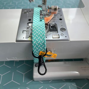 May include: A close-up of a sewing machine with a turquoise and white patterned fabric strip being sewn. The fabric strip has a black metal clip attached to it.