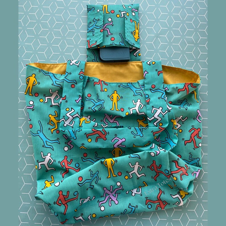 May include: A teal blue reusable tote bag with a yellow lining. The bag features a repeating pattern of colorful stick figures running and playing with a ball. The bag has a small matching pocket on the front.