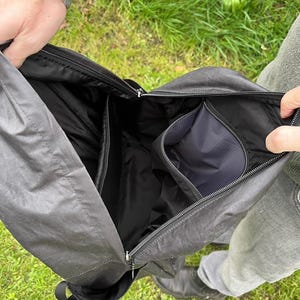May include: Open black messenger bag with multiple internal compartments and a zipped pocket. The bag is made of a durable, water-resistant material. The bag is held open, revealing the interior design.