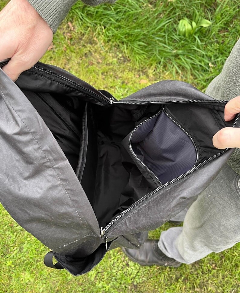 May include: Open black messenger bag with multiple internal compartments and a zipped pocket. The bag is made of a durable, water-resistant material. The bag is held open, revealing the interior design.