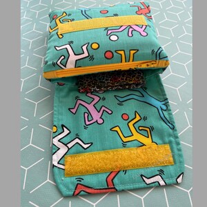 May include: A teal zippered pouch with a yellow zipper and velcro closure. The pouch is decorated with a repeating pattern of colorful stick figures playing with a ball.