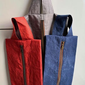 May include: A distinctive tote bag design with a central grey section and a zip, with a red and a blue panel on either side, each with a matching zip. The bag has a pointed top and integrated straps.
