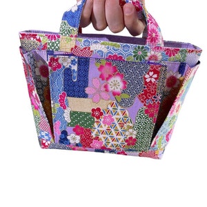 Könnte beinhalten: Eine kleine, violette Tragetasche mit Patchwork-Design, das florale und geometrische Muster in Rosa-, Blau-, Grün- und Goldtönen zeigt. Die Tasche hat einen einzigen Henkel und eine breite, offene Oberseite.