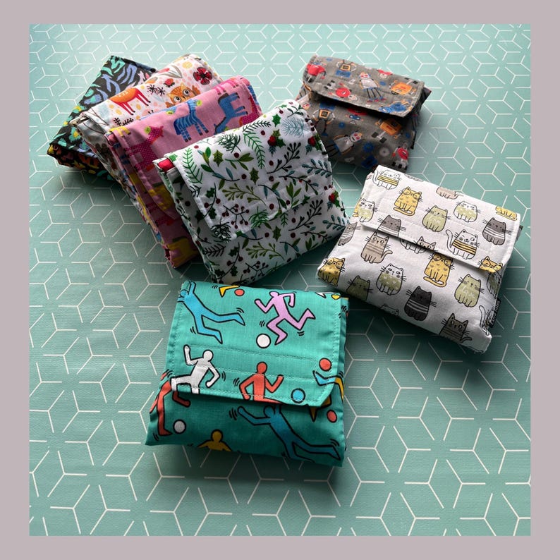 May include: A collection of eight reusable shopping bags made of fabric. The bags are folded and feature different patterns, including a floral design, a cat pattern, a geometric pattern, and a pattern with stick figures.