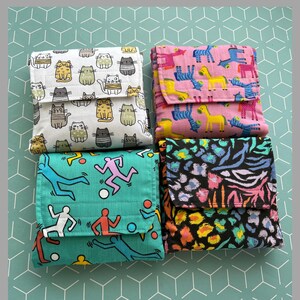 May include: Four reusable shopping bags with different patterns. The first bag has a white background with a repeating pattern of cats. The second bag has a pink background with a repeating pattern of zebras and yellow giraffes. The third bag has a teal background with a repeating pattern of stick figures running. The fourth bag has a black background with a repeating pattern of colorful animal prints.