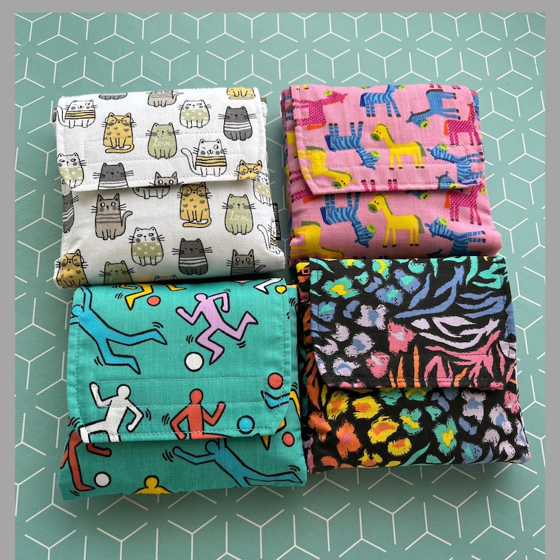 May include: Four reusable shopping bags with different patterns. The first bag has a white background with a repeating pattern of cats. The second bag has a pink background with a repeating pattern of zebras and yellow giraffes. The third bag has a teal background with a repeating pattern of stick figures running. The fourth bag has a black background with a repeating pattern of colorful animal prints.