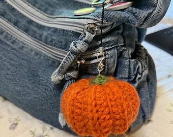 May include: A denim handbag with multiple zippers and a small, orange crocheted pumpkin charm. The pumpkin charm has a green stem and is attached to the bag with a silver clasp. The bag is sitting on a floral patterned surface.