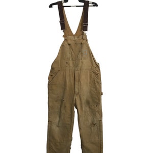 May include: Tan work overalls with brown straps, pockets, and a Carhartt logo patch. The overalls show signs of wear and tear, including holes and patches. The garment is hanging on a black hanger.