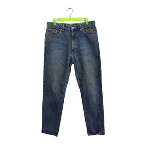 May include: Blue denim jeans hanging on a green hanger. The jeans feature a classic five-pocket design, a button and zip closure, and a faded wash. There are some darker marks on the front of the jeans.