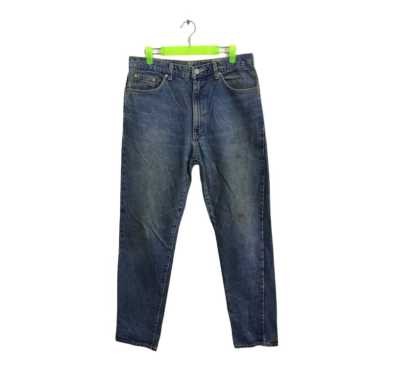 May include: Blue denim jeans hanging on a green hanger. The jeans feature a classic five-pocket design, a button and zip closure, and a faded wash. There are some darker marks on the front of the jeans.