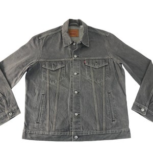 May include: Grey denim jacket with button fastenings, chest pockets, and side pockets. The jacket features a classic collar and a small red tag on the chest pocket. A casual outerwear garment.