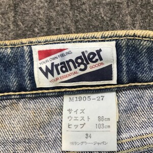 May include: Close-up of a Wrangler jeans label. The label reads "YOUR OWN FEELING. Wrangler GOODS. YOUR ESSENTIAL." A size tag shows measurements: waist 86cm, hips 103cm, and size 34. The jeans are blue denim.