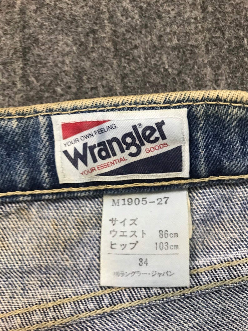 May include: Close-up of a Wrangler jeans label. The label reads "YOUR OWN FEELING. Wrangler GOODS. YOUR ESSENTIAL." A size tag shows measurements: waist 86cm, hips 103cm, and size 34. The jeans are blue denim.