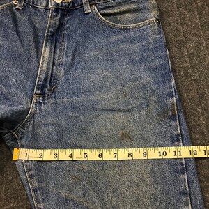 May include: Blue denim jeans with a button closure and a classic five-pocket design. A yellow measuring tape shows a length of approximately 33 cm. The jeans have a faded appearance with some discoloration.