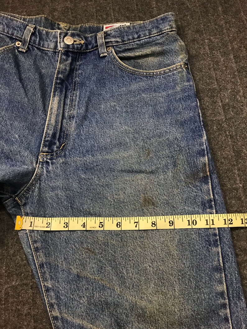 May include: Blue denim jeans with a button closure and a classic five-pocket design. A yellow measuring tape shows a length of approximately 33 cm. The jeans have a faded appearance with some discoloration.