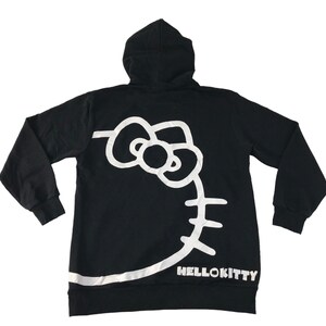May include: Black hooded sweatshirt featuring a large white Hello Kitty graphic on the back. The design includes the iconic bow, face outline, and the text "HELLOKITTY" at the bottom. The sweatshirt has long sleeves and a hood.