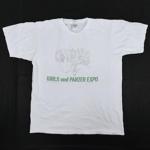 May include: White t-shirt featuring a side profile illustration of an anime character with colorful hair. The text "GIRLS und PANZER EXPO" is printed in green below the illustration. The shirt is made of a soft material.