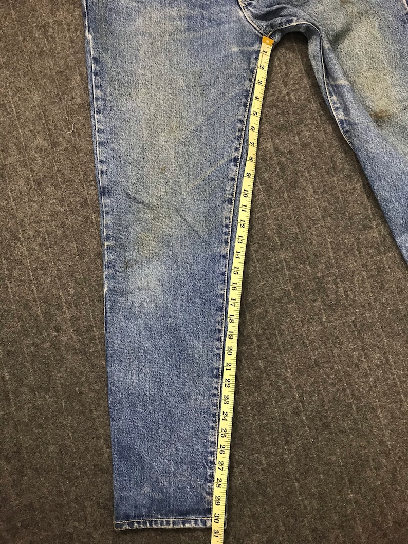 May include: Light blue denim jeans with a measuring tape alongside the leg. The tape indicates a length of approximately 79 cm. The jeans have a classic five-pocket design and a faded wash.