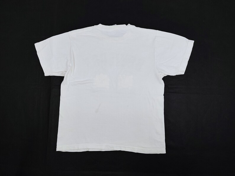 Puede incluir: Una camiseta blanca lisa, vista desde la espalda, con mangas cortas y cuello redondo. La camiseta est&aacute; hecha de una tela suave y opaca y est&aacute; colocada sobre una superficie negra. La apariencia general es simple y casual.