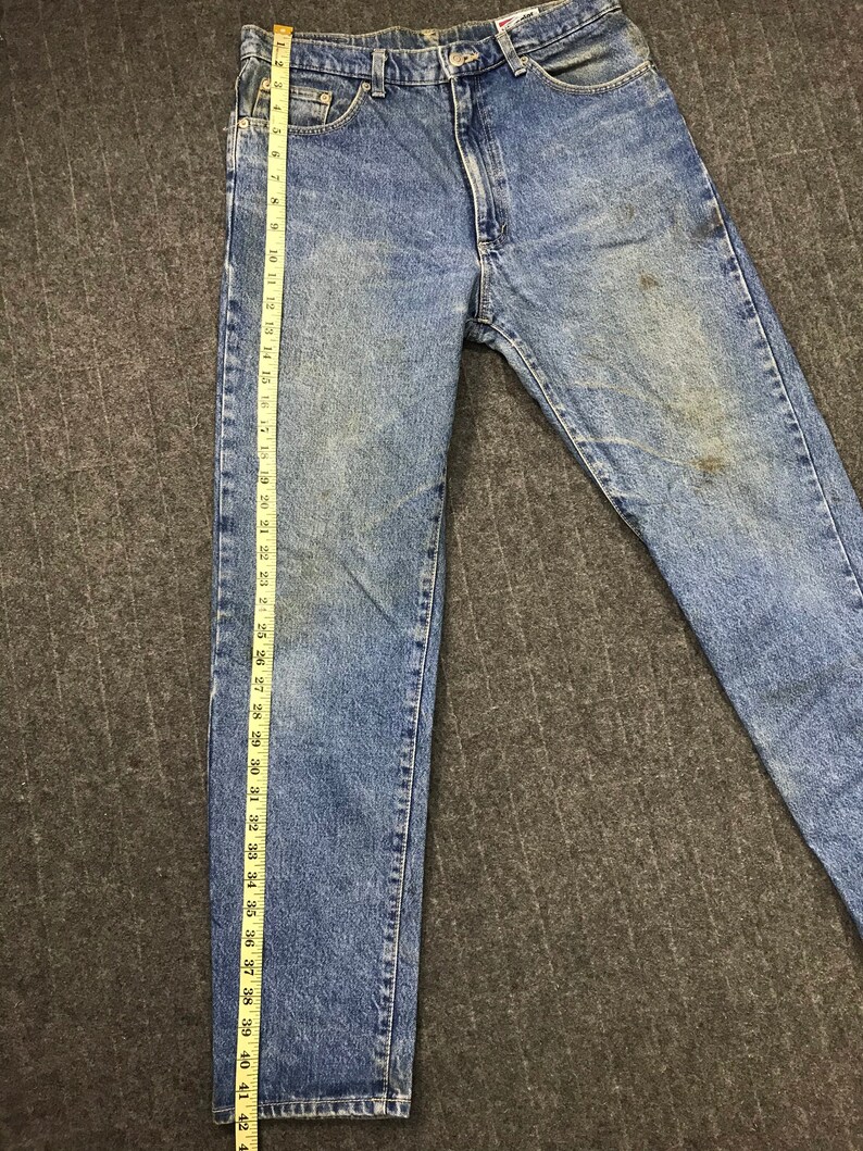 May include: Light blue denim jeans with a straight leg cut. The jeans are measured with a ruler, showing a length of approximately 104 cm. The denim has a faded wash and some discoloration.