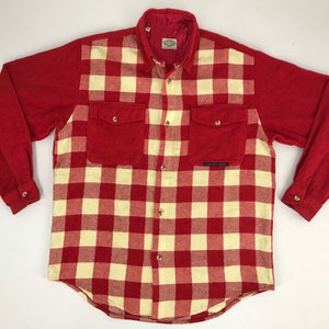 Puede incluir: Una camisa de manga larga con botones que presenta un patrón de cuadros rojos y crema en el panel frontal, mangas rojas sólidas y un cuello rojo sólido. La camisa incluye dos bolsillos en el pecho con solapas abotonadas y cierre central.