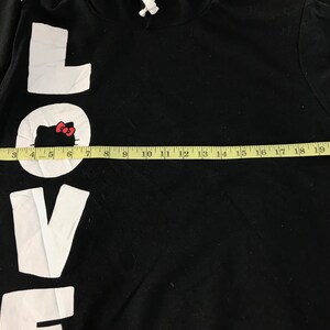 May include: Black hooded sweatshirt with the word "LOVE" in large white letters. The "O" in "LOVE" features a white circle with a Hello Kitty graphic. A yellow measuring tape is laid across the front of the sweatshirt.