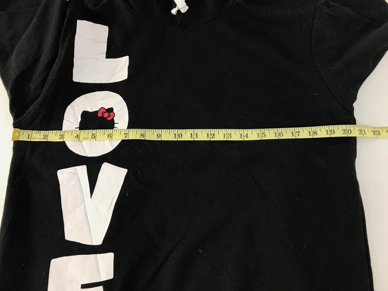 May include: Black hooded sweatshirt with the word "LOVE" in large white letters. The "O" in "LOVE" features a white circle with a Hello Kitty graphic. A yellow measuring tape is laid across the front of the sweatshirt.