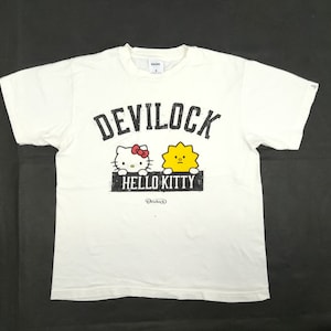 Puede incluir: Camiseta blanca con la inscripción "DEVILOCK" sobre un gráfico de Hello Kitty y un personaje amarillo con forma de sol. Las palabras "HELLO KITTY" están impresas debajo de los personajes. La camiseta tiene cuello redondo y mangas cortas.
