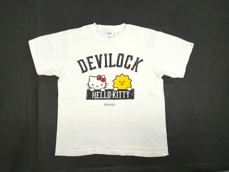 Puede incluir: Camiseta blanca con la inscripci&oacute;n "DEVILOCK" sobre un gr&aacute;fico de Hello Kitty y un personaje amarillo con forma de sol. Las palabras "HELLO KITTY" est&aacute;n impresas debajo de los personajes. La camiseta tiene cuello redondo y mangas cortas.