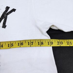 Puede incluir: Camiseta blanca con un dise&ntilde;o gr&aacute;fico negro. Una cinta m&eacute;trica amarilla muestra el ancho de la camiseta. La camiseta est&aacute; hecha de un material suave y c&oacute;modo.