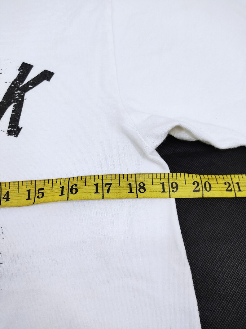 Puede incluir: Camiseta blanca con un dise&ntilde;o gr&aacute;fico negro. Una cinta m&eacute;trica amarilla muestra el ancho de la camiseta. La camiseta est&aacute; hecha de un material suave y c&oacute;modo.