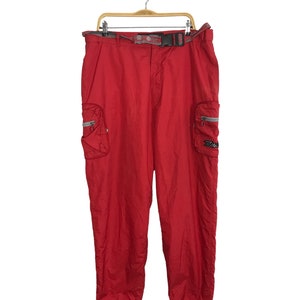 Könnte beinhalten: Rote Cargohose mit passendem Gürtel, hängend an einem Holzbügel. Die Hose hat zwei Seitentaschen mit Reißverschlüssen und ein kleines Logo am rechten Bein. Die Hose ist aus leichtem Material.